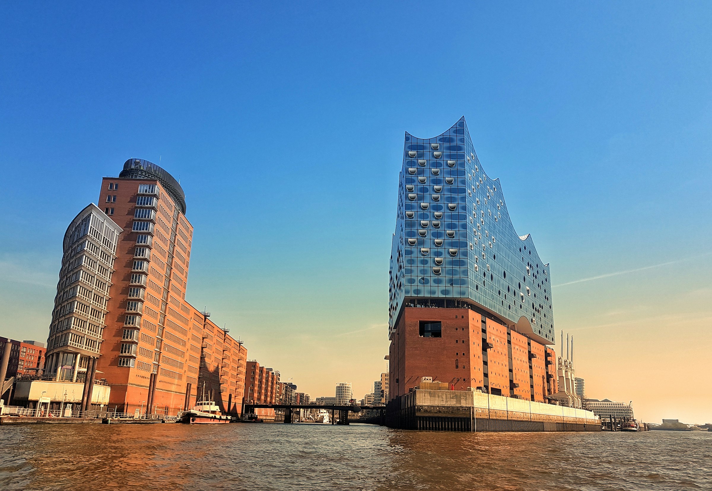 The glass wave-topped Elbphilharmonie on a brick warehouse base, beside a tall brick office tower, over the water.