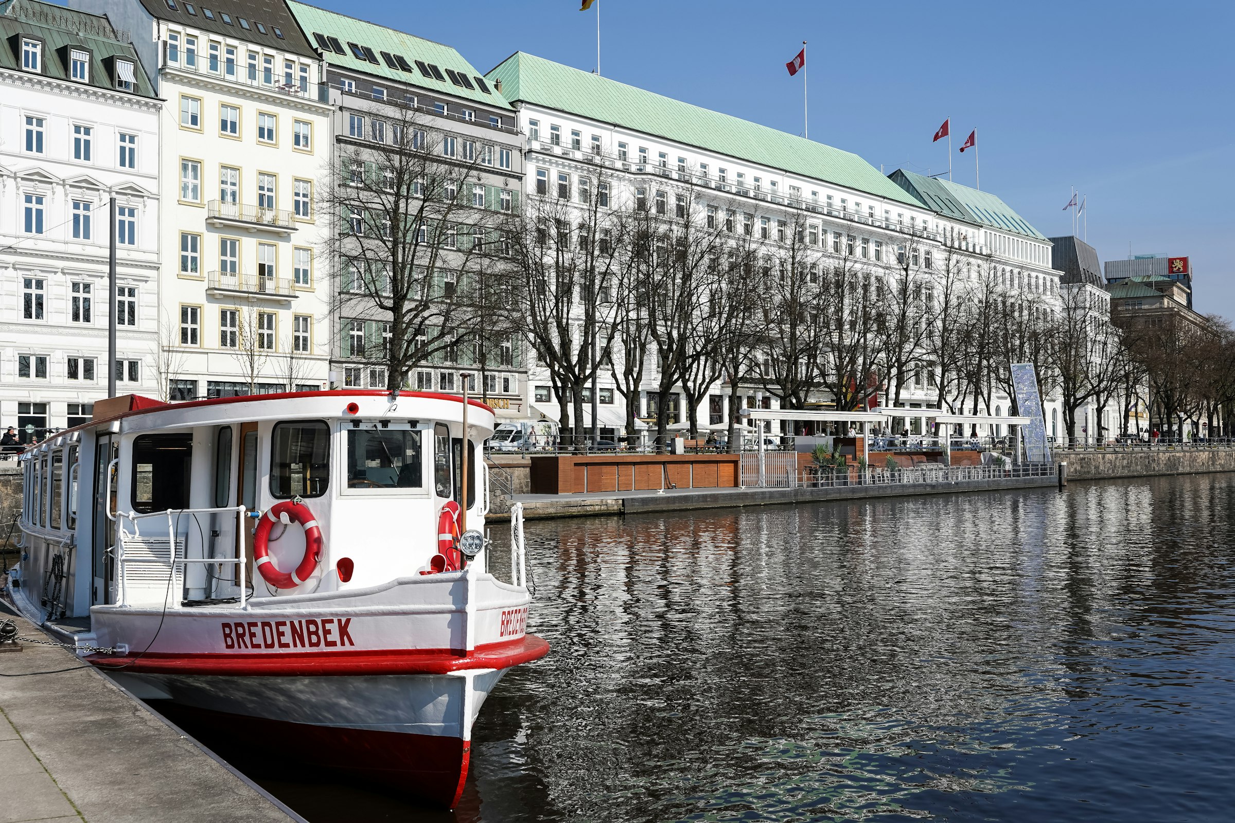 A white Alster ferry moored by a promenade, with grand white hotel façades and bare trees behind.