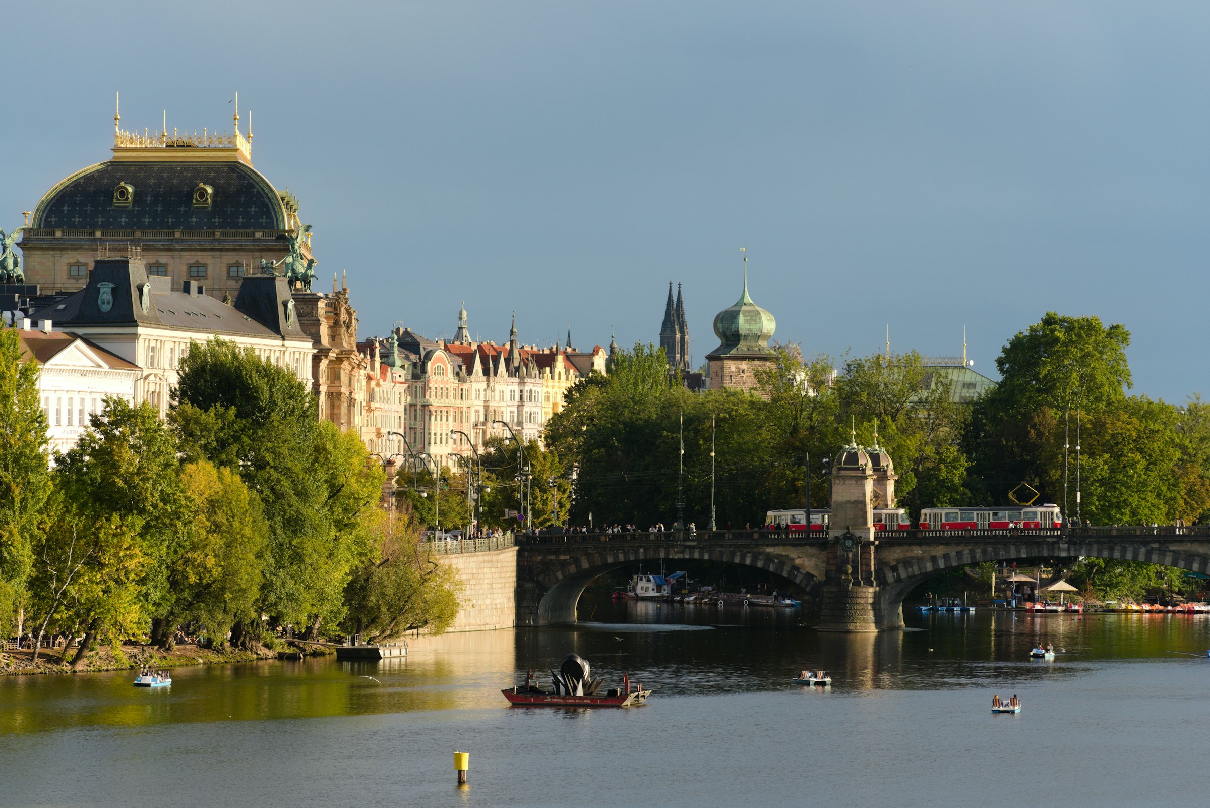 The gold-domed National Theatre above trees on the riverbank, with a tram crossing a stone bridge.