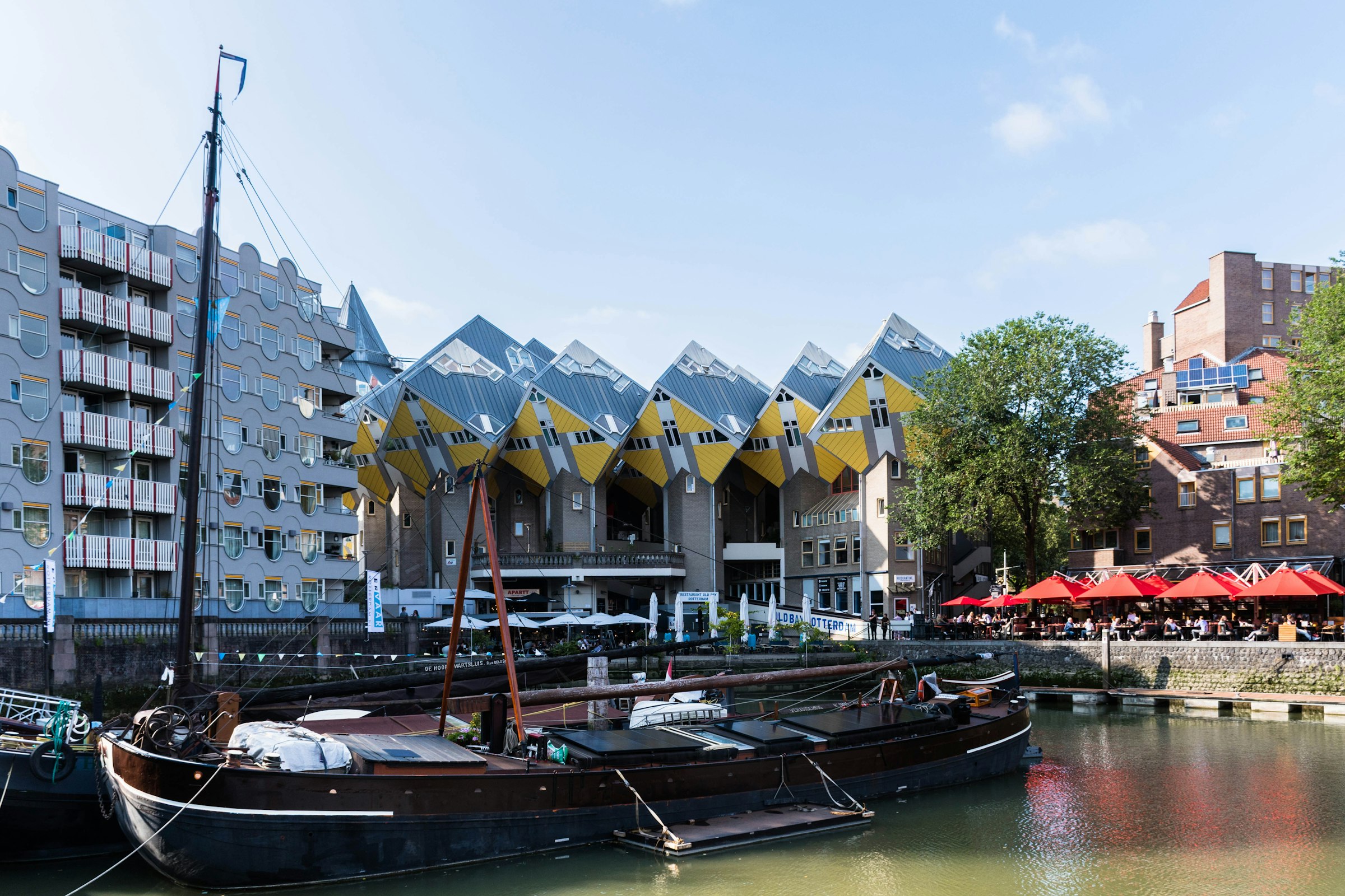 Old sailing barges moored in a small harbour, with yellow tilted cube houses and café umbrellas behind.
