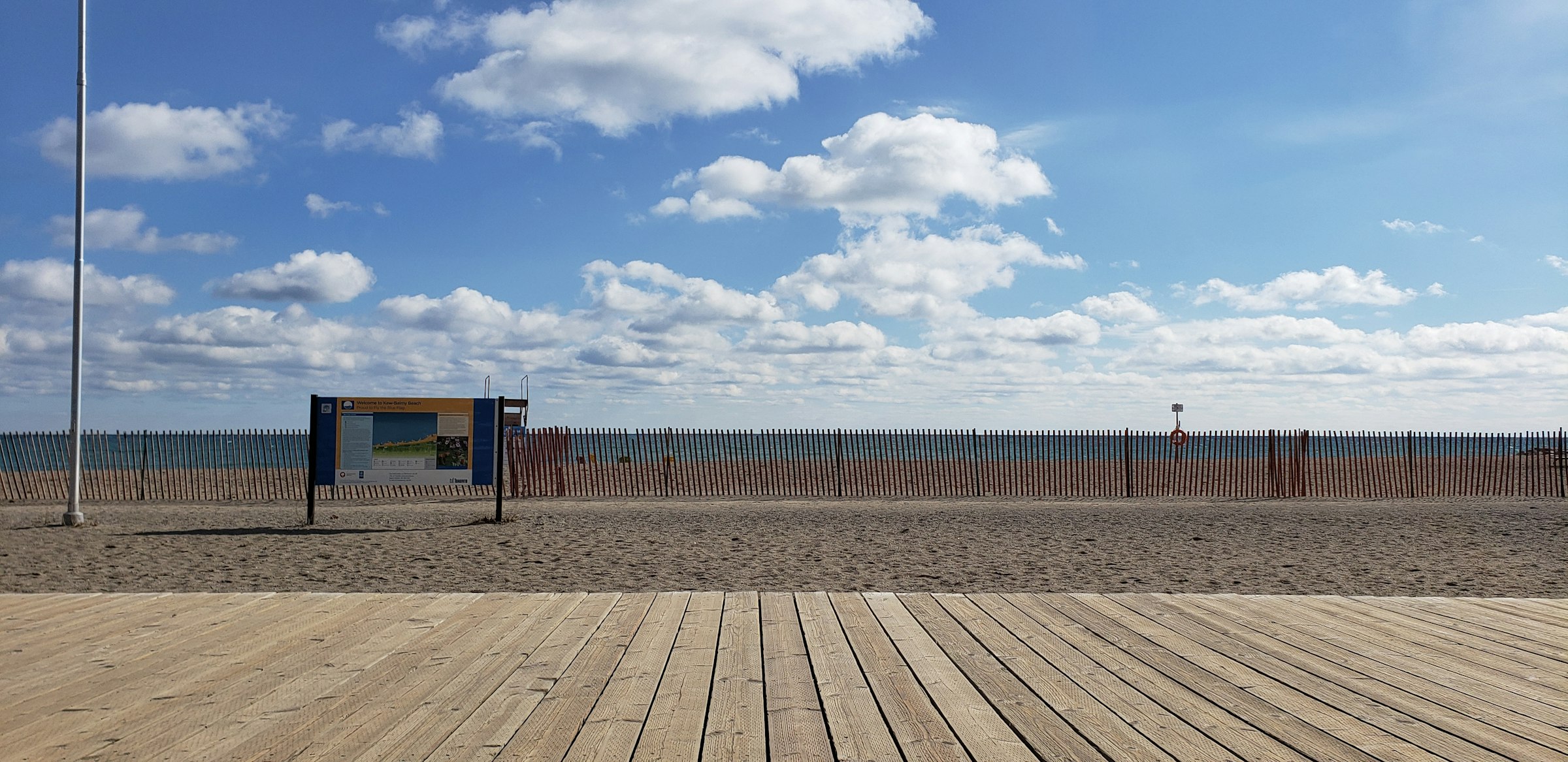 An empty wooden boardwalk beside a wide sand beach on Lake Ontario under a blue sky.